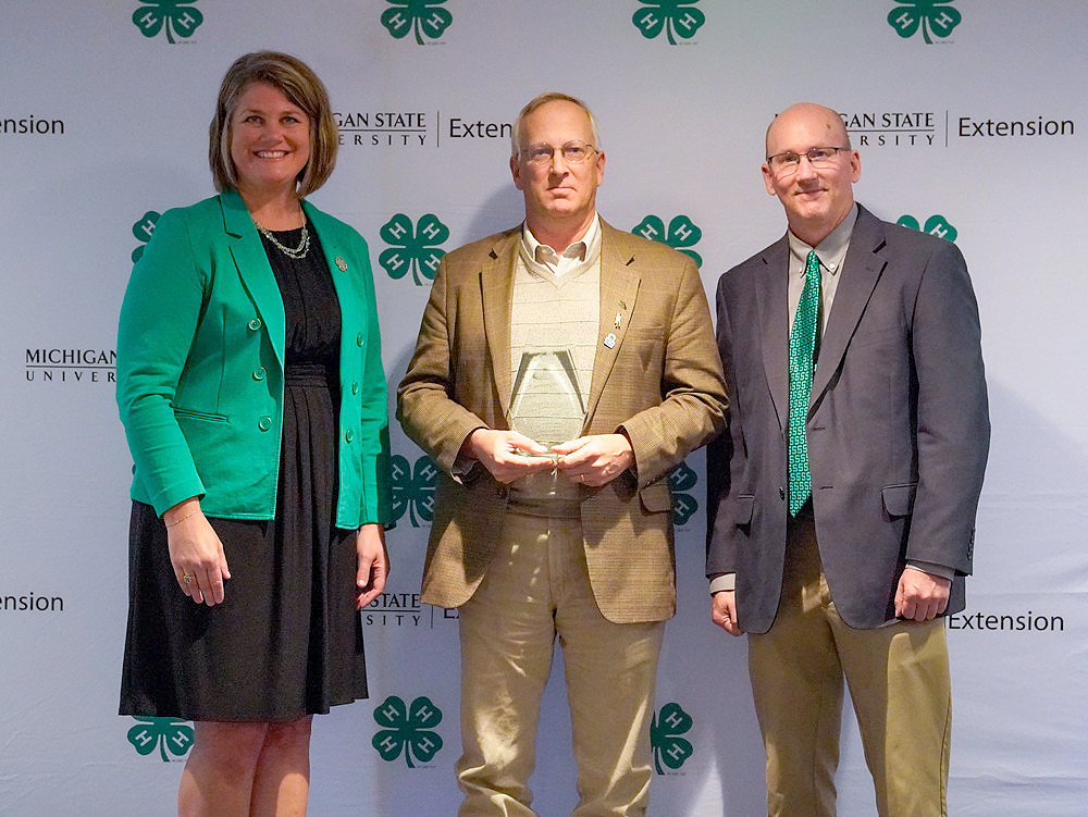 Three individuals at a backdrop. The center person holds an award.
